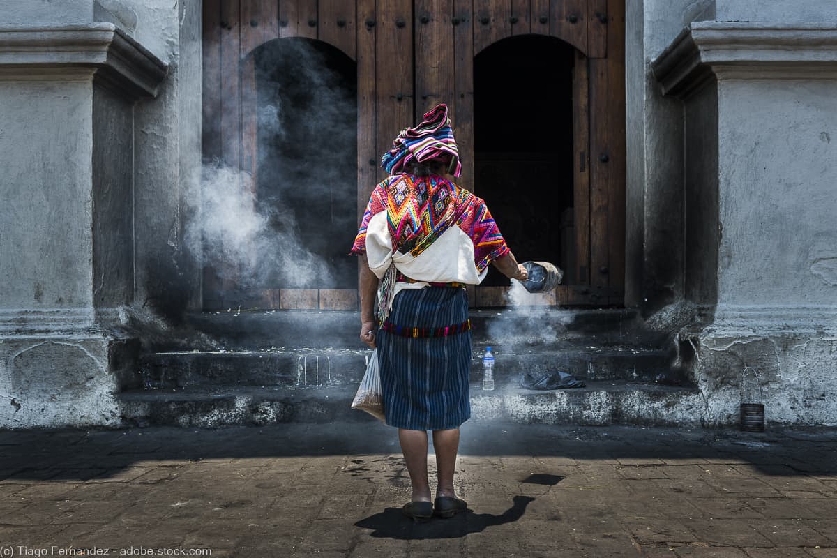 the back of an elderly Maya woman standing in front of a church and waving incense around