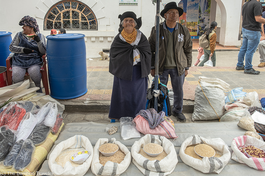 An elderly woman and man standing in front of bags of rice and dried corn at a market in Otavalo