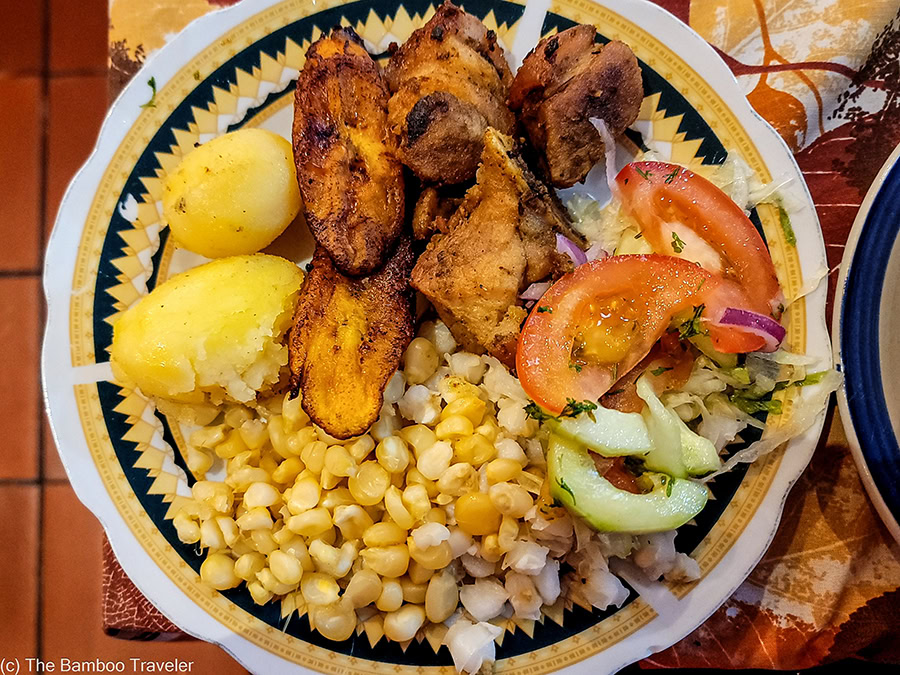 A plate of traditional Ecuadorian fritada consisting of fried pork chunks, sweet fried plantains, boiled potatoes, and a mountain of mote (hominy corn). A side salad of tomato, onion, and cucumber sits on the edge of the patterned ceramic plate.