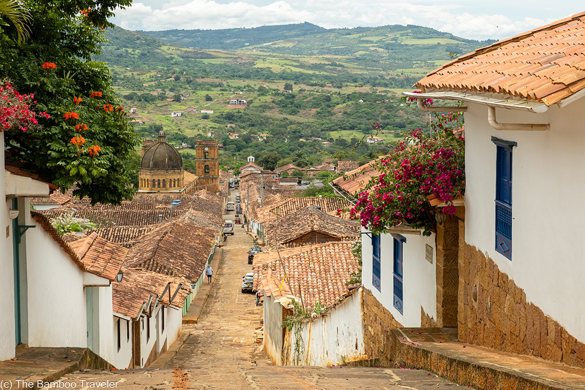 a cobblestoned street going down hill lined with whitewash adobe single story buildings with red tile roofs and green mountains in the distance