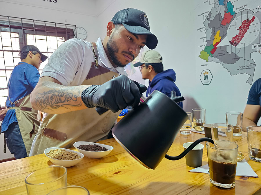A professional barista in black gloves carefully pouring hot water from a gooseneck kettle into a glass during a specialty coffee tasting at a Divino Coffee Workshop Bogota tour.