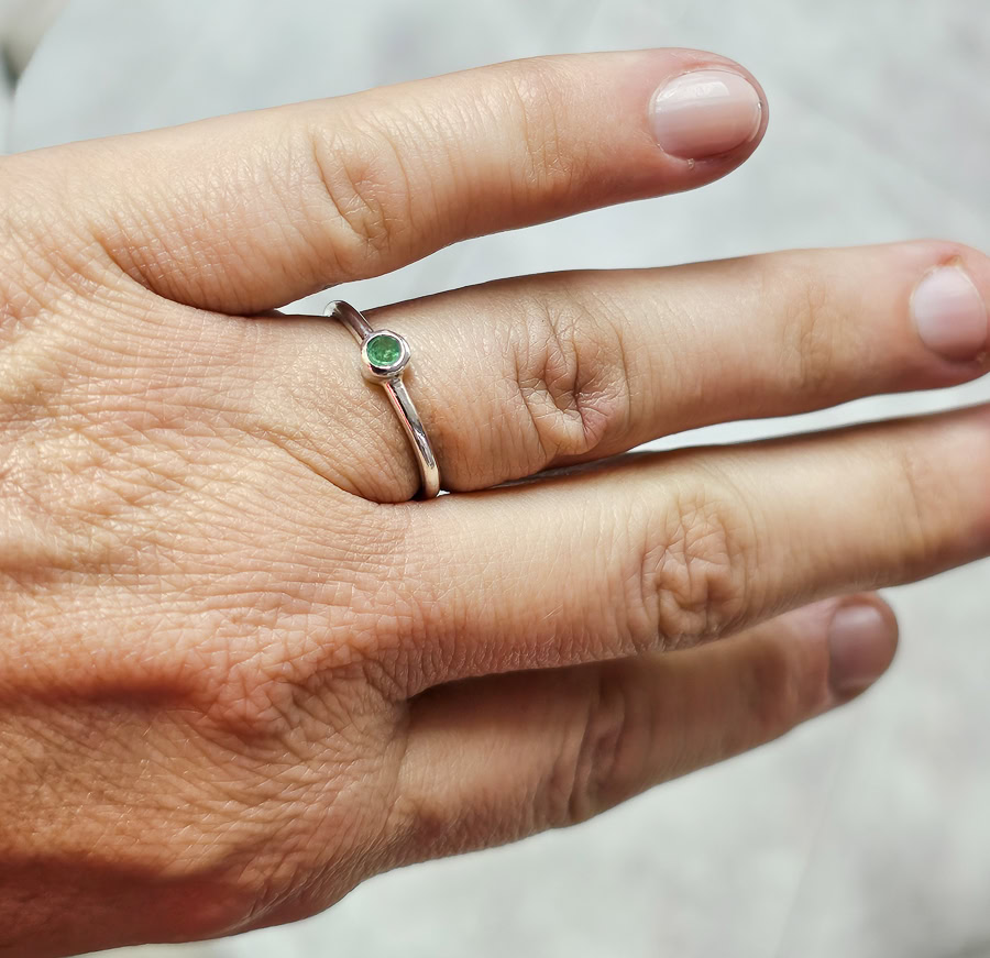 Close-up of a person's hand wearing a thin silver band with a single, small green emerald set in a bezel. The ring is worn on the ring finger against a neutral, out-of-focus background.