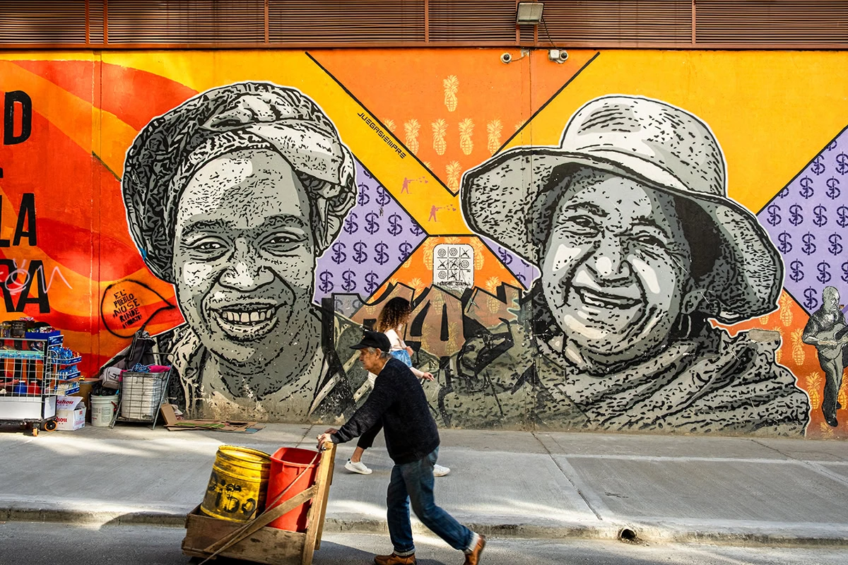 A local vendor walks past a massive, vibrant graffiti mural of Afro-Colombian women during a guided street art tour in Bogota