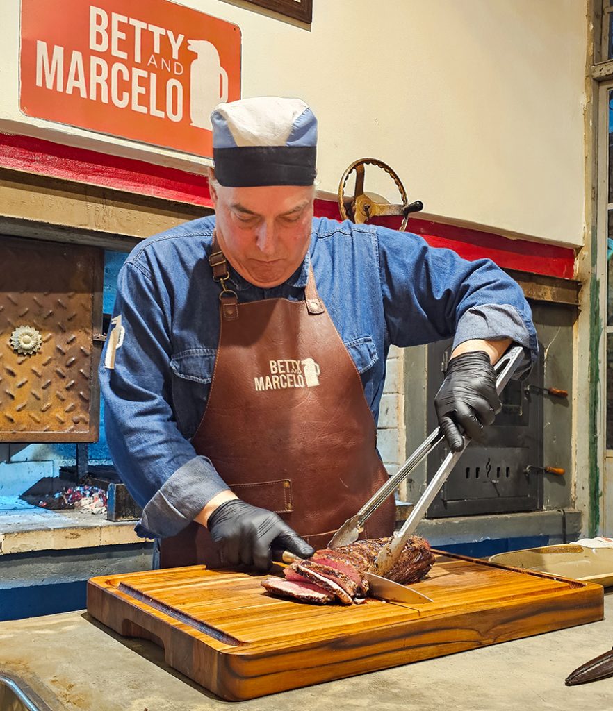 Host Marcelo wearing a branded leather apron and black gloves as he carefully carves a colita de cuadril (tri-tip steak) on a wooden cutting board. 