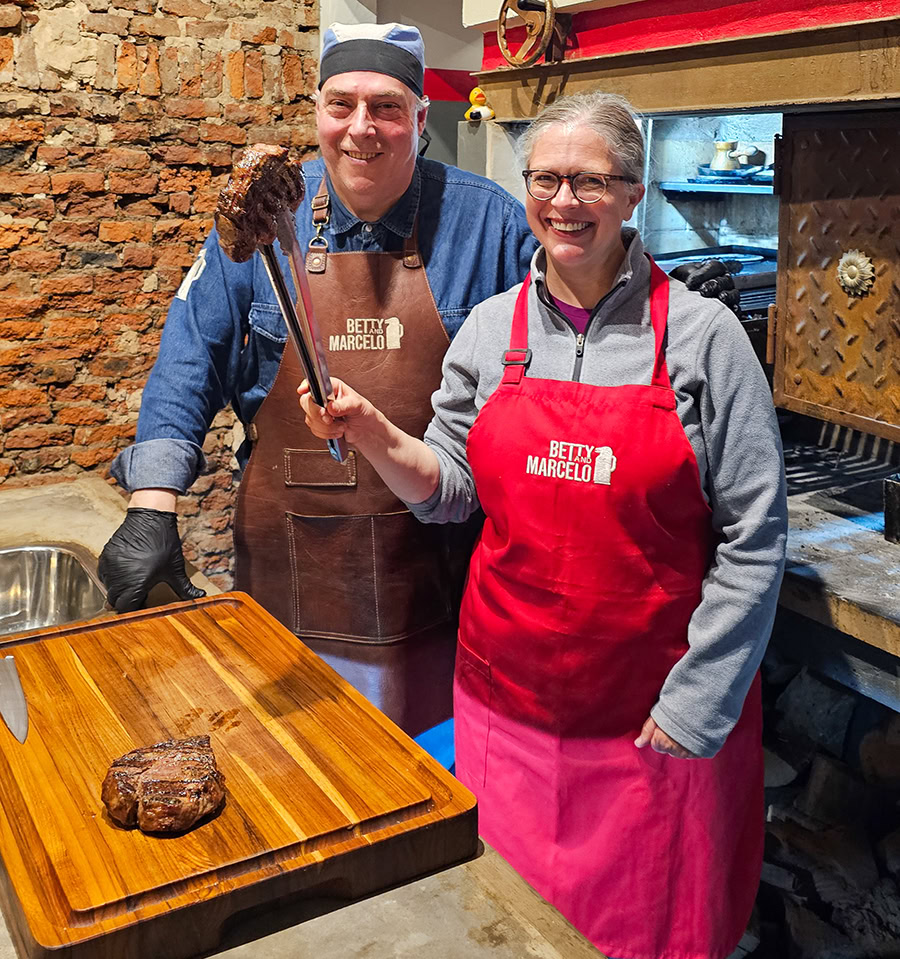One person wearing a red apron and holding a piece of meat with tongs and standing next to a man in a brown apron and a cap