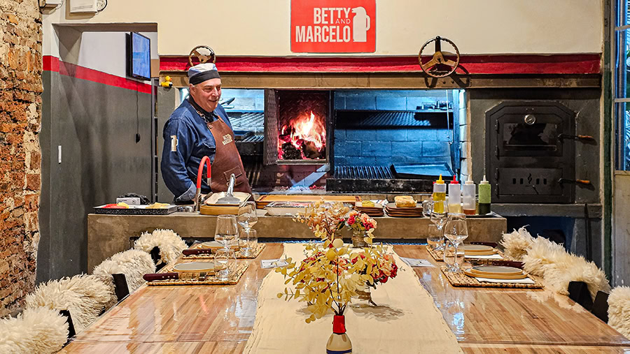 A man wearing a blue shirt and a brown leather apron stands behind a counter in a rustic kitchen. He is smiling and looking toward a large indoor grill area where a bright wood fire is burning in a central hearth. The counter in front of him is set with various ingredients, condiments, and cooking tools. In the foreground, a dining table is elegantly set with woven placemats, wine glasses, floral arrangements, and chairs draped with white sheepskin. A red and white sign on the wall above the grill reads "BETTY AND MARCELO."