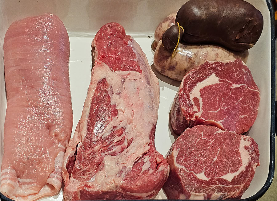 A top-down view of a white tray featuring various cuts of raw Argentine beef and traditional sausages for an asado in Buenos Aires. The selection includes a large matambre (flank steak), thick-cut ojo de bife (ribeye), a lean roast, and traditional chorizo and morcilla (blood sausage). This represents the high-quality, local ingredients used during Betty and Marcelo's Asado Food Tour in Argentina.