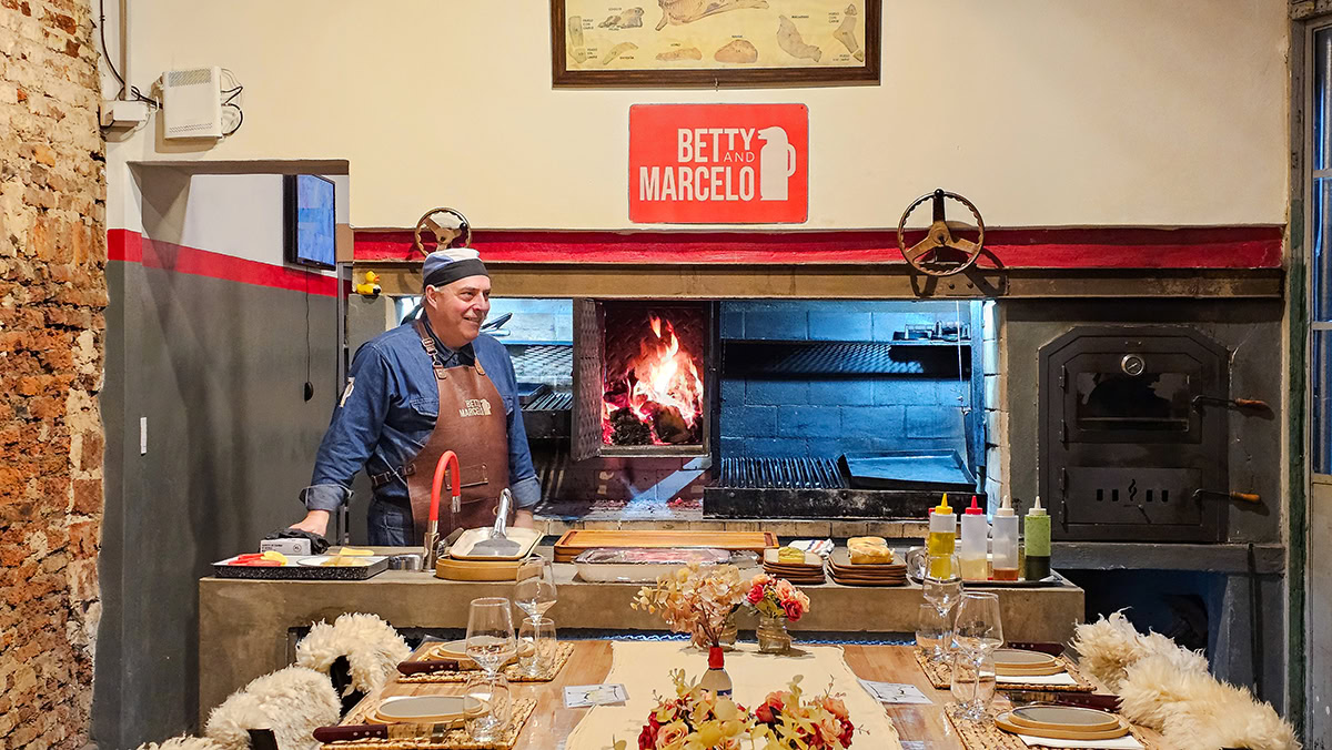 A man wearing a blue shirt and a brown leather apron stands behind a counter in a rustic kitchen. He is smiling and looking toward a large indoor grill area where a bright wood fire is burning in a central hearth. The counter in front of him is set with various ingredients, condiments, and cooking tools. In the foreground, a dining table is elegantly set with woven placemats, wine glasses, floral arrangements, and chairs draped with white sheepskin. A red and white sign on the wall above the grill reads "BETTY AND MARCELO."