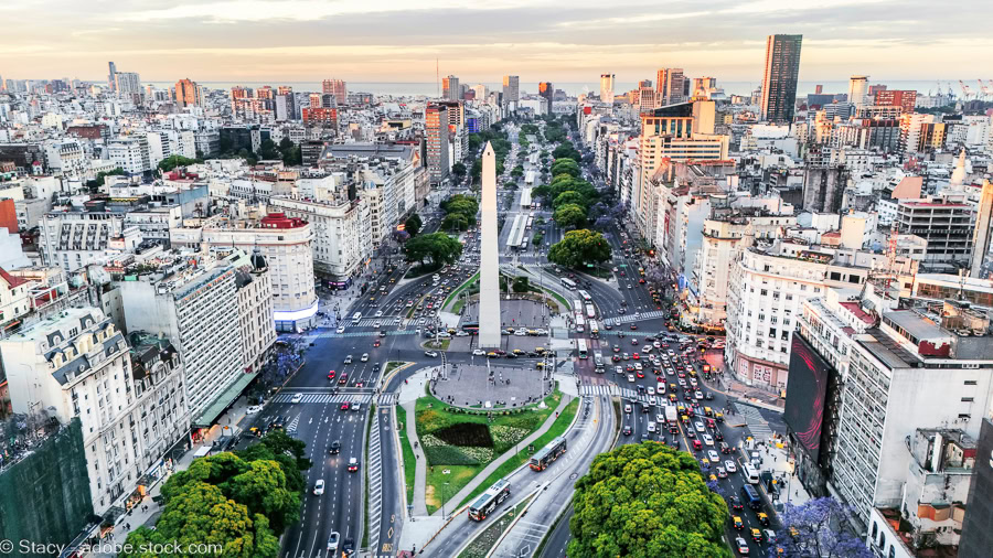 An aerial, high-angle view of the Obelisco de Buenos Aires standing at the center of the massive Avenida 9 de Julio. The image shows heavy city traffic, lush green trees lining the wide boulevards, and the sprawling urban skyline of the Argentine capital during the soft light of golden hour.