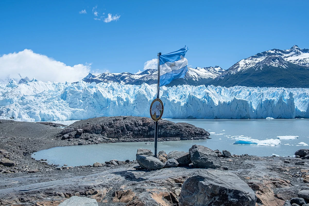 A view of the Perito Moreno Glacier on the Blue Safari tour, with the Argentine flag waving in the foreground against a clear blue sky, icebergs, and the vast glacier wall.