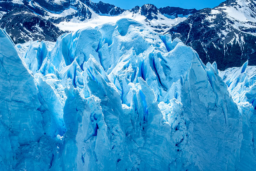 A close-up view of the towering, fractured blue and white ice formations of the Perito Moreno Glacier, contrasting against the dark, snow-dusted Southern Andes mountains in the background.