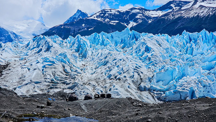 A field of dark rocks and gravel leads to the rugged base of the Perito Moreno Glacier, contrasting dirty-white and deep-blue ice with small wooden huts along the shore under a mountain backdrop.