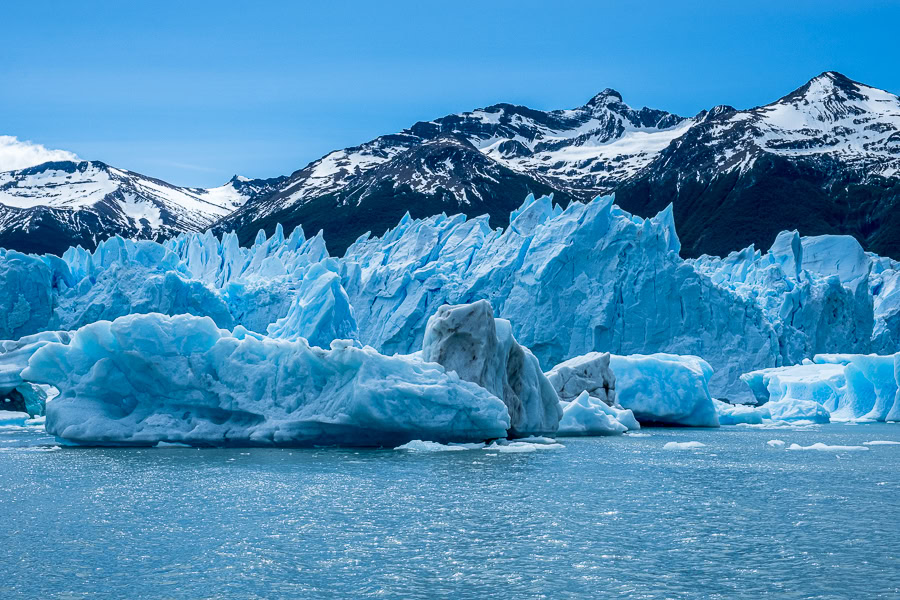 The massive, serrated edge of the Perito Moreno Glacier showing deep blue crevasses and floating ice chunks in the gray-blue water during the Blue Safari boat tour.