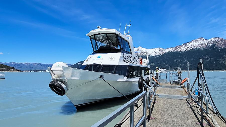 A white sightseeing catamaran docked at the Bajo de las Sombras port on the turquoise waters of Lago Argentino, with snow-capped Andes mountains in the background.