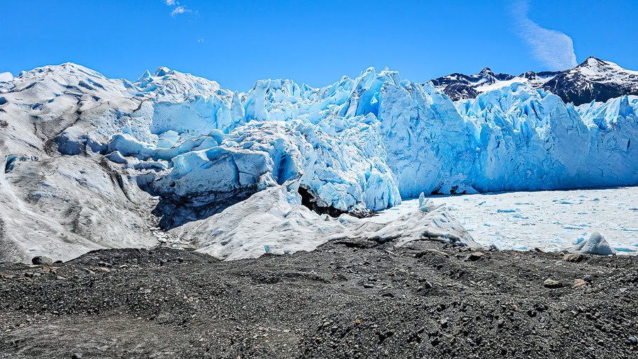 Wide view of the edge of the Perito Moreno Glacier, contrasting a debris field of dark rocks and gravel with jagged, dirty-white and bright-blue ice walls under a clear sky.