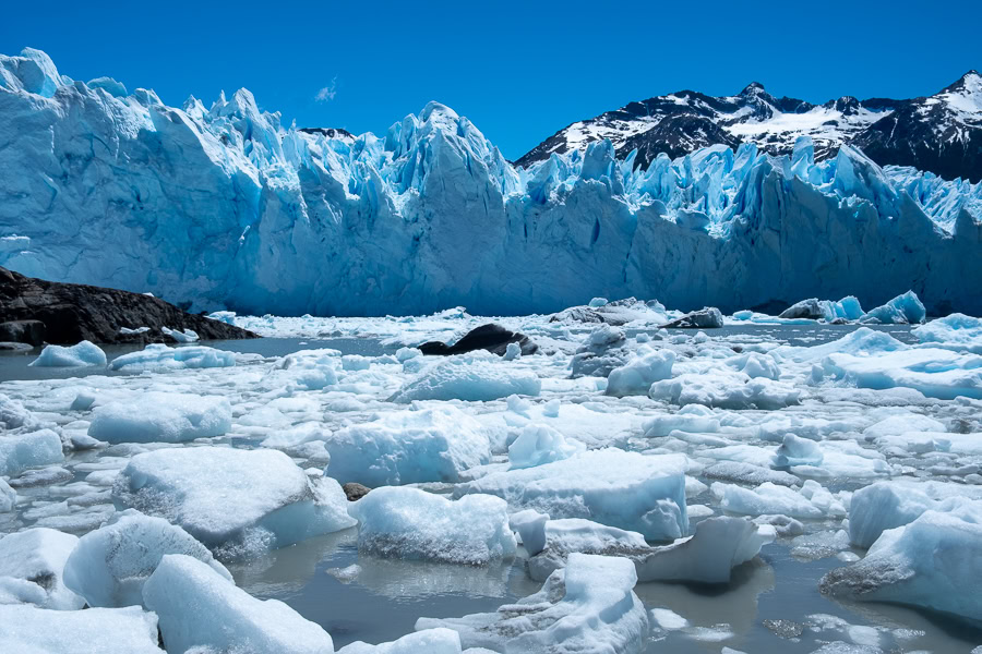A field of floating ice chunks fills the foreground of Lago Argentino, leading to the massive, bright blue vertical wall of the Perito Moreno Glacier, with snow-capped mountains and a blue sky in the background.