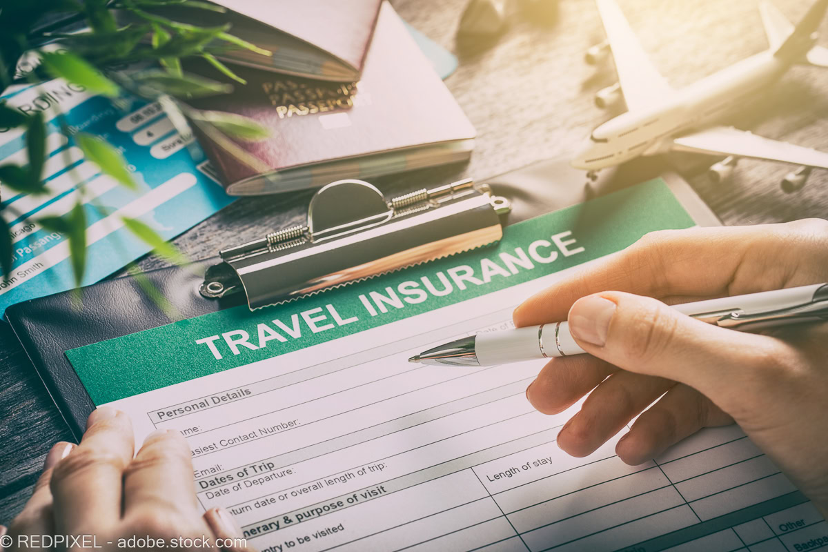 A close-up, top-down shot of a person’s hands holding a pen over a paper travel insurance application form on a clipboard. In the background, out-of-focus travel items like a passport, boarding pass, and a small toy airplane are visible on a wooden surface under warm light.