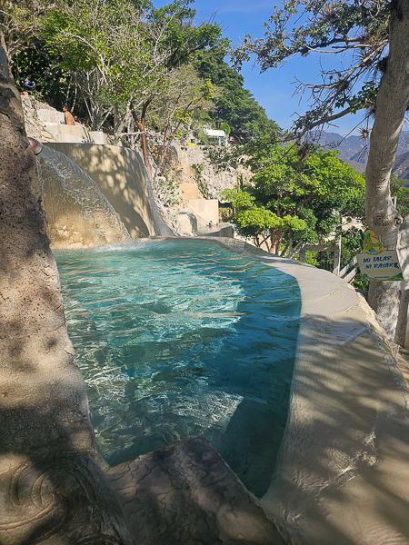 A close-up view of a thermal pool at Grutas Tolantongo in Hidalgo, Mexico, showing warm spring water cascading from an upper level into the clear, turquoise tub.