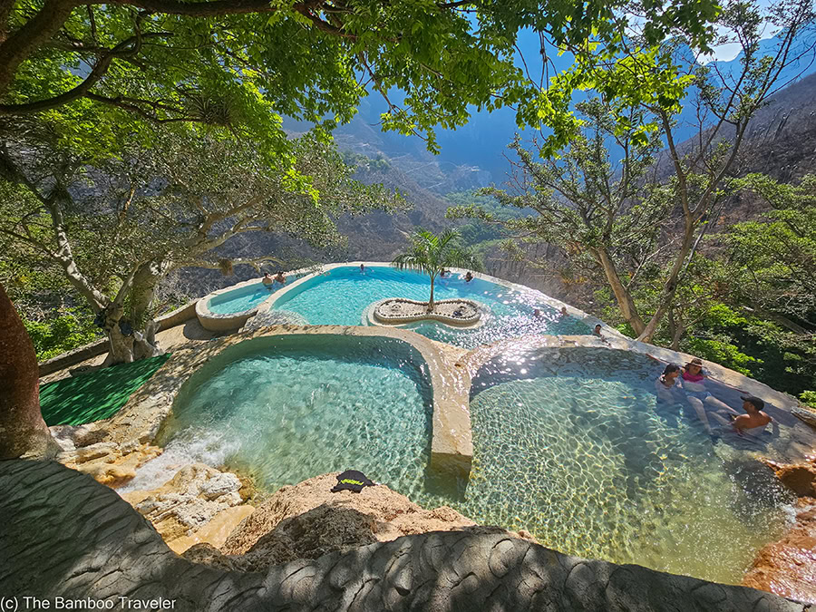 tourists relaxing in three crystal-clear thermal pools at Grutas Tolantongo in Hidalgo, Mexico, featuring a small palm tree island with a dramatic canyon drop-off in the background.