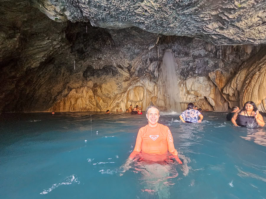 Julie standing in the warm, turquoise thermal pool inside the main cave at Grutas Tolantongo in Hidalgo, Mexico, with water cascading down the rock wall behind her.