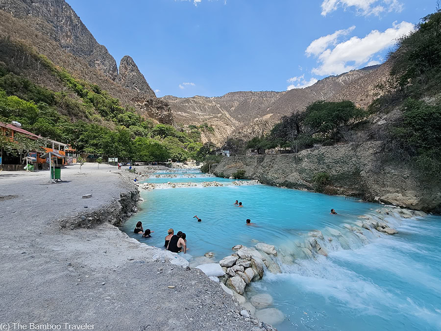 A wide-angle landscape shot of the bright turquoise Tolantongo River flowing through a deep canyon with tourists swimming in the thermal pools and rugged mountains under a blue sky.