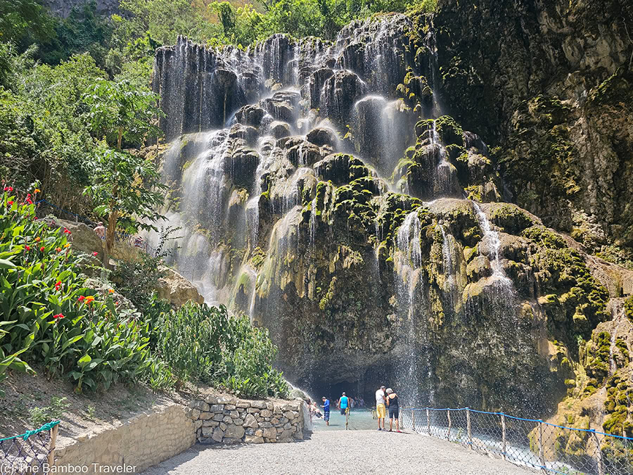 A daytime photo of the main waterfall at Grutas Tolantongo in Hidalgo, Mexico, showing water cascading down a large, mossy cave entrance. A few tourists are standing at the entrance to the cave.