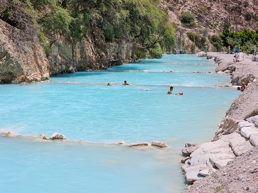 A daytime shot of the tranquil, bright turquoise Tolantongo River flowing through a rugged canyon. Six tourists are swimming and wading in the shallow thermal water, which is sectioned by low stone barriers.